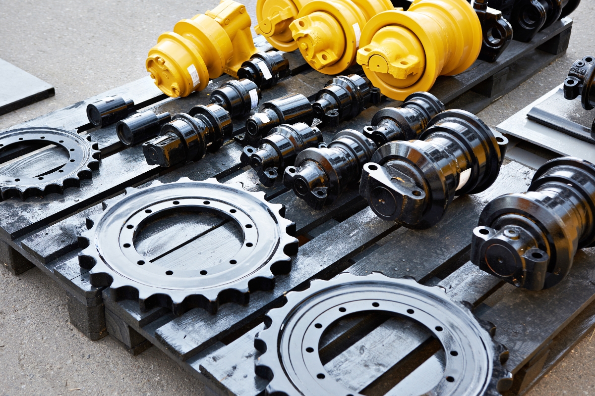 Close-up of large, yellow hydraulic components and black metal sprockets and rollers for heavy machinery laid out on a wooden pallet.